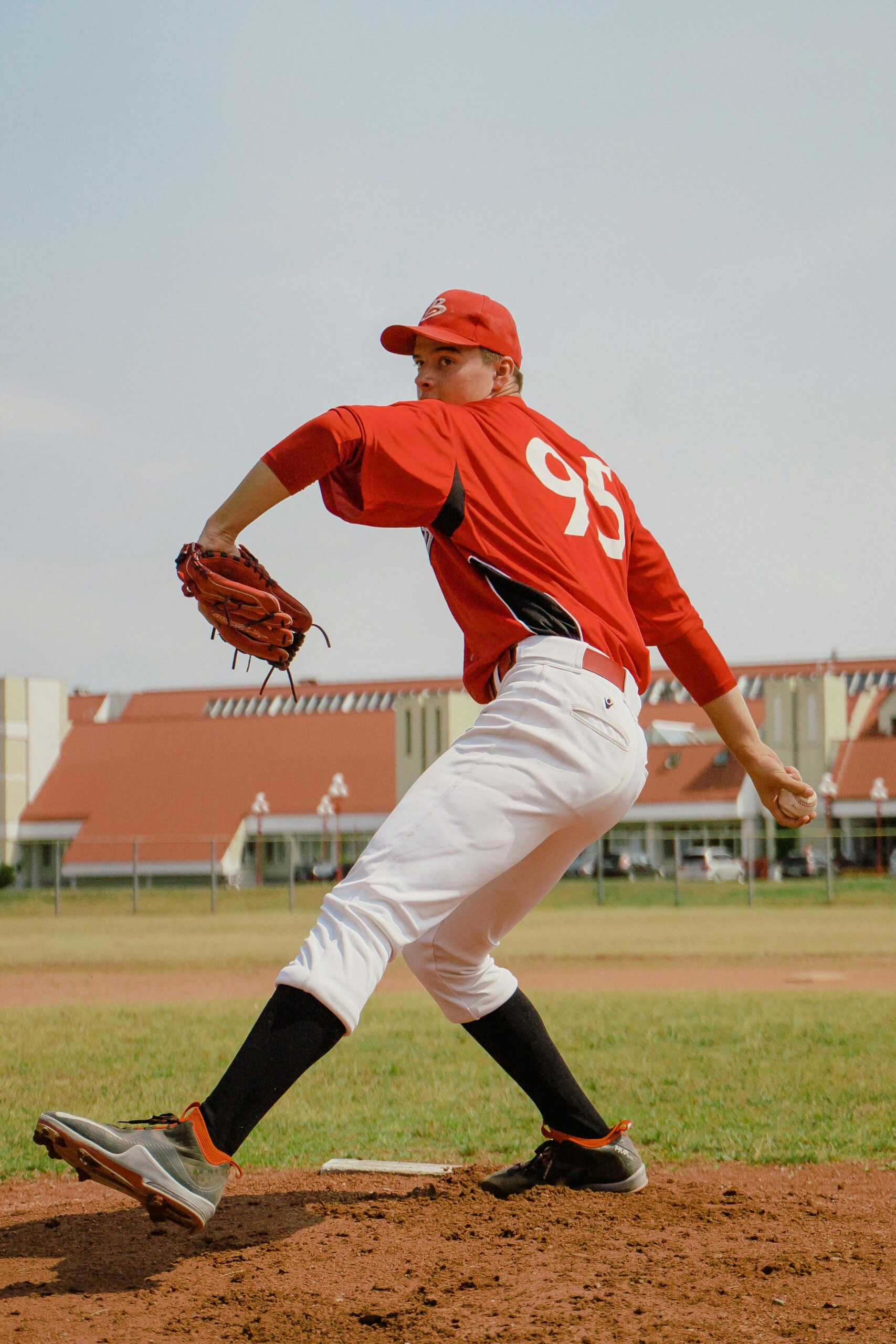 A baseball pitcher in a red uniform with the number 95 winding up to throw a pitch on a grassy field, with buildings in the background.